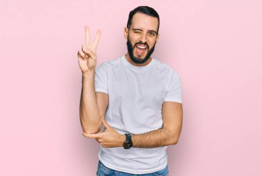 Sakallı, sıradan beyaz tişörtlü, gülümseyen ve kameraya göz kırpan genç bir adam parmaklarıyla zafer işareti yapıyor. İki numara.. 