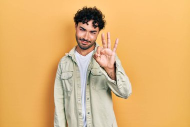 Gündelik kıyafetler giyen genç İspanyol adam üç numarayı gösterip işaret ederken kendinden emin ve mutlu gülümsüyor.. 