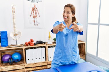 Orta yaşlı İspanyol fizyoterapist kadın. Ağrı kesici kliniğinde çalışıyor. Seni ve kamerayı işaret ediyor. 