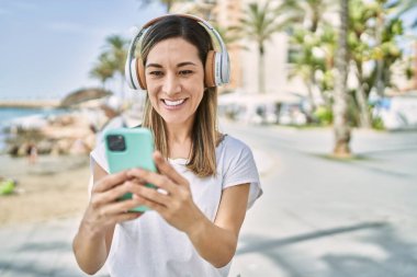 Genç İspanyol kadın deniz kenarında akıllı telefon ve kulaklık kullanarak gülümsüyor.