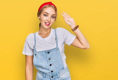 Gündelik giysiler giymiş genç beyaz kız merhaba diyor mutlu ve gülümsüyor, dostça karşılama hareketi yapıyor. 