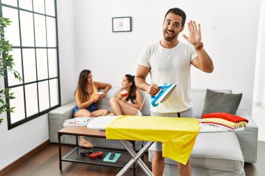 Genç İspanyol adam evde ütü yapıyor merhaba diyor mutlu ve gülümsüyor, dostça karşılama hareketi yapıyor. 