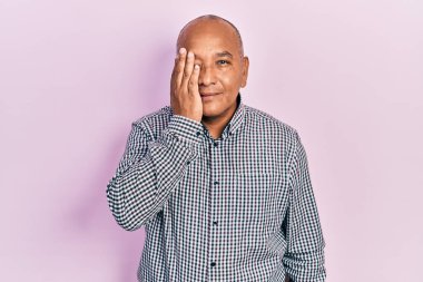 Orta yaşlı, sıradan kıyafetler giyen, bir gözünü eliyle kapatan, yüzünde kendine güvenen bir gülümseme ve sürpriz duygular taşıyan Latin bir adam.. 