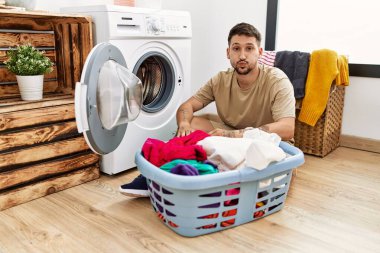 Young handsome man putting dirty laundry into washing machine puffing cheeks with funny face. mouth inflated with air, crazy expression. 