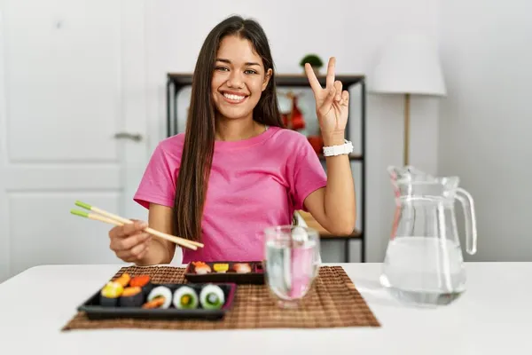 年轻的黑发女人在吃寿司时 用筷子微笑着看着摄像机 展示手指在做胜利的标志 第二点 — 图库照片