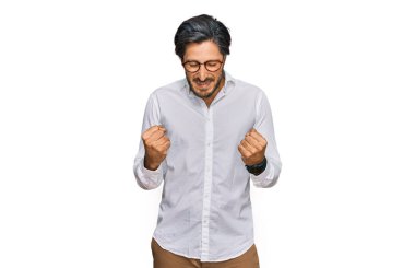 İş gömleği ve gözlük takan genç İspanyol adam çok mutlu ve heyecanlı. Kolları havada, gülümseyen ve başarı için bağıran kazanan bir jest yapıyor. kutlama konsepti. 