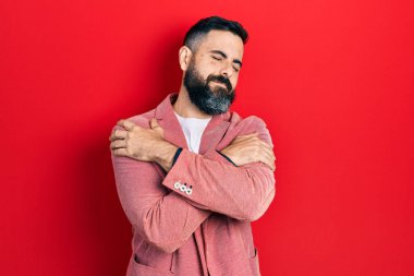 Latin kökenli genç bir adam iş kıyafetleri giyip kendine sarılıyor mutlu ve pozitif, kendinden emin gülümsüyor. Sevgi ve özgüven. 