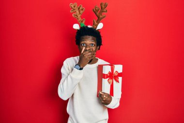 Geyik Noel şapkası takan genç Afro-Amerikan bir adam elinde hediyeyle ağzını yanlışlıkla ellerle kapatırken şok geçirdi. gizli kavram. 