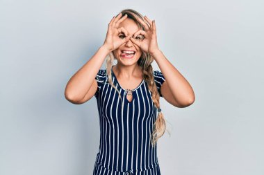 Güzel, genç, sarışın bir kadın, sıradan çizgili bir elbise giyiyor, dürbün gibi jest yapıyor, dili dışarı çıkarıyor, gözleri parmaklarından bakıyor. çılgın ifade. 