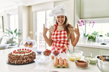 Güzel, genç, esmer pasta şefi kadın mutfakta pasta pişirirken çılgın bir ifadeyle elleri havada rock sembolü yapıyor. Müzik yıldızı. ağır kavram. 
