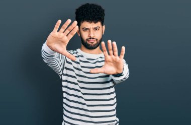 Sakallı, sıradan çizgili süveter giyen, el ve parmaklarını kullanarak çerçeve yapan genç bir Arap. 