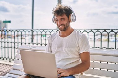 Genç İspanyol adam deniz kenarında dizüstü bilgisayar kullanarak gülümsüyor.