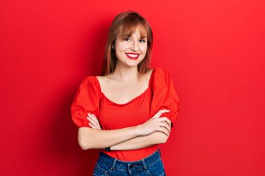 Kırmızı tişörtlü, mutlu yüzlü, kollarını kavuşturup kameraya bakan kızıl saçlı genç kadın. pozitif kişi. 