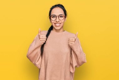 Gündelik kıyafetler ve gözlük takan genç İspanyol kız. Başarı işareti ile el hareketi yapıyor. Gülümsüyor ve mutlu. Neşeli ifade ve kazanan jest. 
