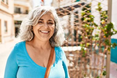 Orta yaşlı, kır saçlı, gülümseyen bir kadın şehirde mutlu mesut duruyor..