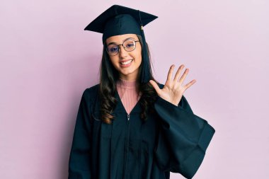 Mezuniyet şapkası ve tören cübbesi takan genç İspanyol kadın beş numarayı gösterip işaret ederken kendinden emin ve mutlu bir şekilde gülümsüyor.. 