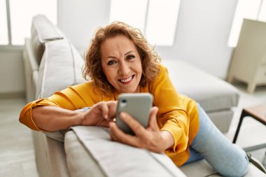 Orta yaşlı beyaz kadın akıllı telefon kullanıyor. Evdeki kanepede oturuyor..