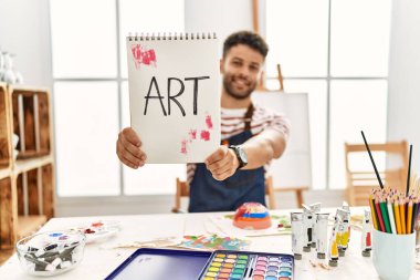 Genç Arap sanatçı gülümsüyor. Sanat stüdyosunda elinde sanat sözcüğüyle mutlu bir not defteri tutuyor..