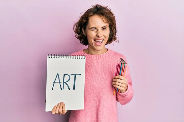Młoda Brunetka Trzyma Notatnik Artystyczny Kolorowe Ołówki Mrugając Patrząc Aparat — Zdjęcie stockowe