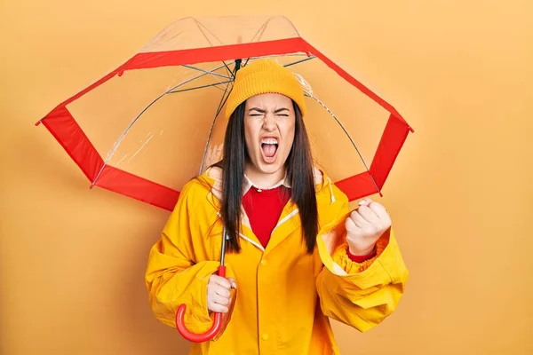 黄色のレインコートを着た若いヒスパニック系の女性が傘を持っている怒りでいらいらし フラストレーションの叫び 怒りと手を上げてクレイジー叫んで — ストック写真