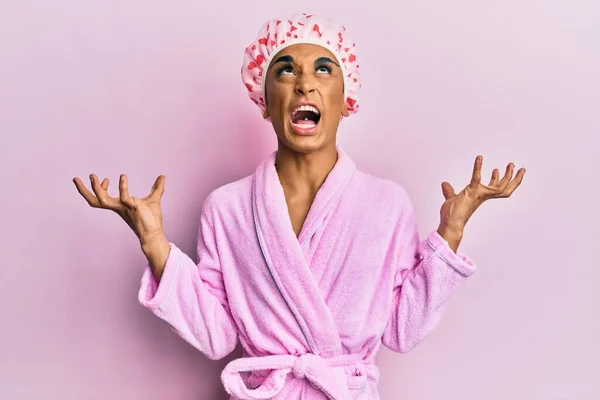Hispanic man wearing make up wearing shower towel cap and bathrobe crazy and mad shouting and yelling with aggressive expression and arms raised. frustration concept.