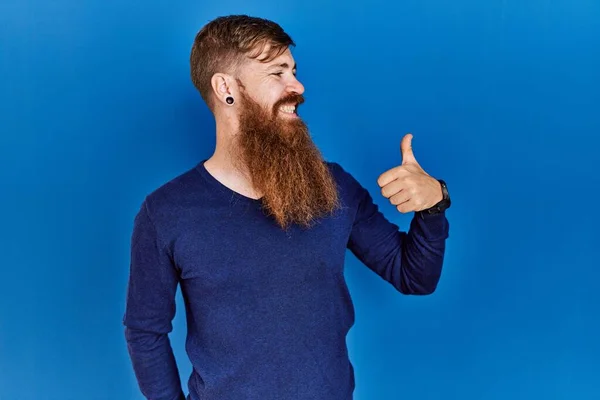红头发的男人留着长胡子 穿着休闲的蓝色毛衣 背景蓝色 看起来很自豪 微笑着向旁边竖起大拇指 — 图库照片
