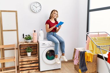 Dokunmatik ped kullanan genç beyaz bir kız evde çamaşır makinesinin başında oturmuş bekliyor..