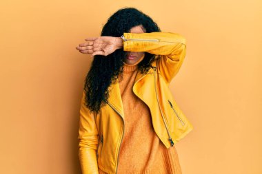 Orta yaşlı Afro-Amerikalı kadın yün kazak ve deri ceket giyip gözlerini kollarıyla kapatıyor, ciddi ve üzgün görünüyor. Görme, saklama ve reddetme konsepti 