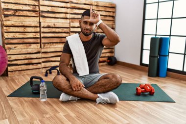 Spor salonundaki antrenman minderinde oturan genç Hintli. Alnında parmakları olan insanlarla dalga geçiyor. Ezik hareketler yapıyor.. 