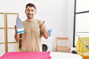 Young handsome man ironing clothes at home smiling happy and positive, thumb up doing excellent and approval sign 