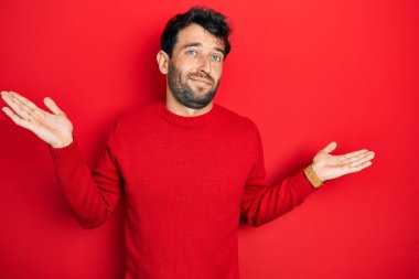 Sakallı, sıradan kırmızı süveter giyen, kafası karışmış, kolları ve elleri havada yakışıklı bir adam. şüphe kavramı. 