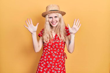 Sarışın, güzel, beyaz bir kadın. Yazlık şapka takıyor ve on numarayı işaret ediyor. Kendinden emin ve mutlu bir şekilde gülümsüyor.. 