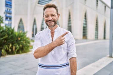 Yüzünde mutlu ve doğal bir ifadeyle, elleri ve parmakları kenarı işaret eden, güler yüzlü orta yaşlı bir adam şehrin dışında. 