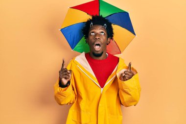 Sarı yağmurluk giyen Afro-Amerikalı genç adam şaşırmış ve şaşırmış bir şekilde yukarı baktı ve parmaklarıyla işaret etti ve kollarını kaldırdı.. 