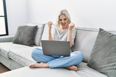 Young caucasian woman using laptop at home sitting on the sofa very happy and excited doing winner gesture with arms raised, smiling and screaming for success. celebration concept. 