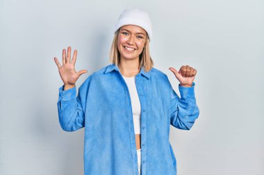 Güzel sarışın kadın yün şapka takıyor ve altı numaralı parmaklarıyla işaret ederken kendinden emin ve mutlu gülümsüyor.. 