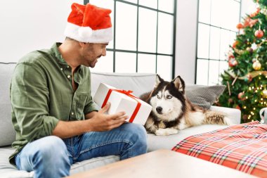 Genç İspanyol bir adam elinde hediyeyle evdeki Noel ağacının yanında köpekle koltukta oturuyor.