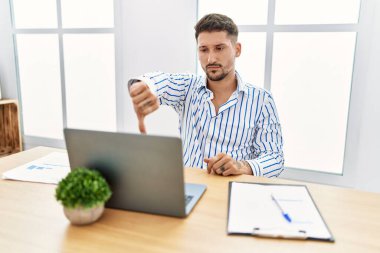 Young handsome man with beard working at the office using computer laptop looking unhappy and angry showing rejection and negative with thumbs down gesture. bad expression. 