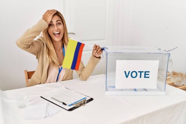 Blonde beautiful young woman at political campaign election holding colombia flag stressed and frustrated with hand on head, surprised and angry face 