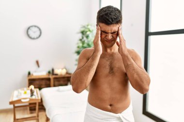 Young hispanic man standing shirtless at spa center with hand on head for pain in head because stress. suffering migraine. 