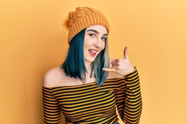 Yün şapka takan, gülümseyen, el ve parmaklarıyla telefon hareketi yapan, telefonda konuşan genç, modern bir kız. iletişim kavramları. 