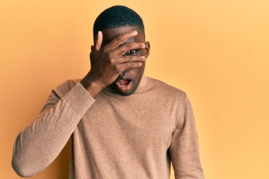 Sıradan bir kazak giyen genç Afro-Amerikalı adam şok içinde yüzünü ve gözlerini eliyle kapatıyor, parmaklarından korkuyla bakıyor. 