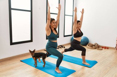 Spor merkezinde köpekle yoga eğitimi alırken gülümseyen iki kadın.