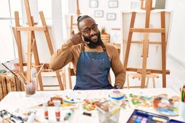 African american artist man at art studio smiling doing phone gesture with hand and fingers like talking on the telephone. communicating concepts. 