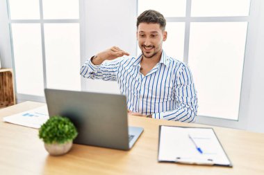 Young handsome man with beard working at the office using computer laptop gesturing with hands showing big and large size sign, measure symbol. smiling looking at the camera. measuring concept. 