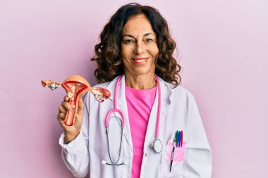 Orta yaşlı İspanyol doktor kadın, kadın üreme organının anatomik modelini tutuyor. Pozitif ve mutlu görünüyor. Kendine güvenen bir gülümsemeyle dişlerini gösteriyor. 