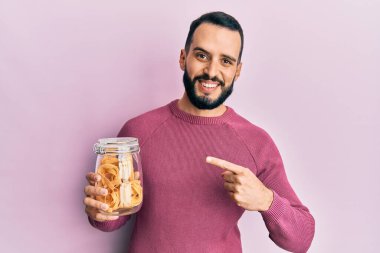 Elinde sakallı, elinde makarna kavanozuyla gülümseyen mutlu eli ve parmağı olan genç bir adam. 