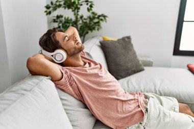 Genç İspanyol adam ellerini başının üstüne koyup evde müzik dinliyor..