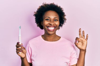 Genç Afro-Amerikalı kadın elinde elektrikli diş fırçası tutuyor parmaklarıyla işaret yapıyor, gülümsüyor ve mükemmel bir sembol oluşturuyor. 