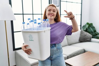 Geri dönüşüm sepetini elinde tutan genç kızıl saçlı kadın plastik şişelerle gülümsüyor ve el hareketleriyle küçük boylarda işaret veriyor parmaklarıyla kameraya bakıyor. ölçüm kavramı. 